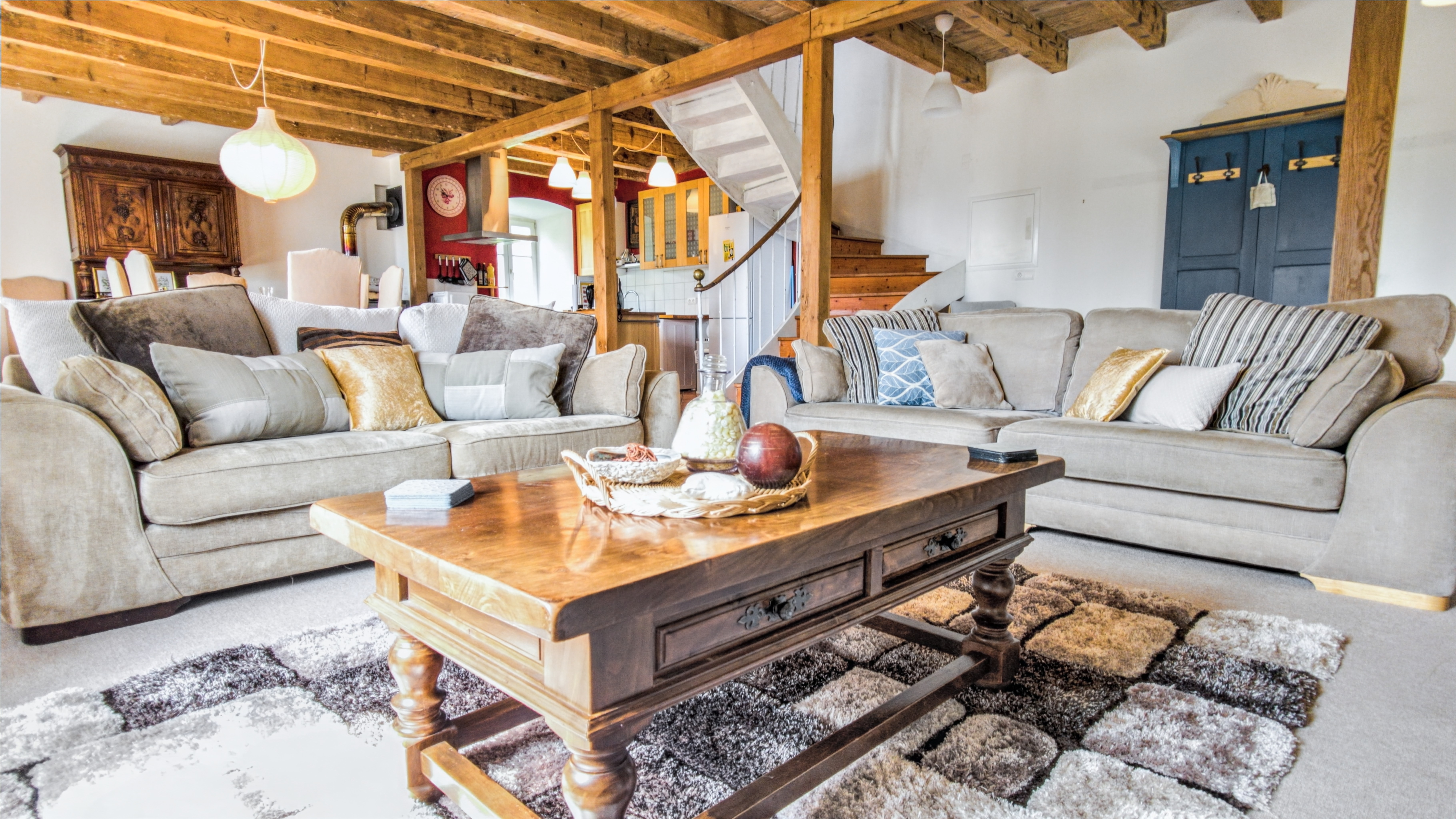 Grand salon chaleureux avec poutres en bois au plafond, comprenant deux grands canapés beige remplis de coussins aux tons [...]